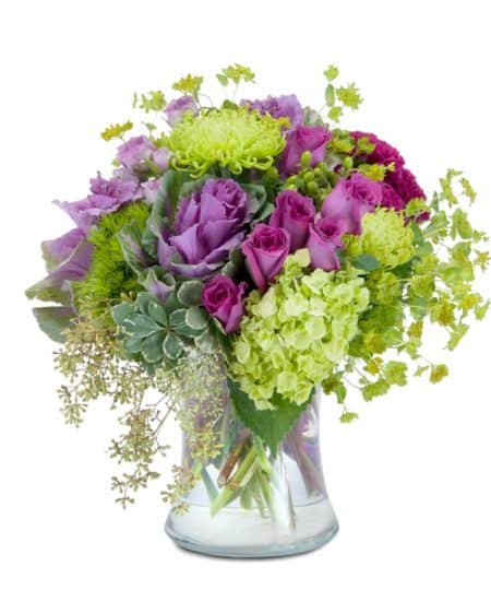 A magical mixture of magenta and green flowers, including roses, hydrangea, hypericum berry and ornamental kale, designed in a large clear glass vase.