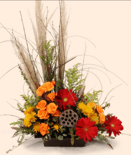 Pampas Grass add softness and height to the bouquet. Wild Grasses add color and texture throughout the one sided design that is perfectly suited for a buffet. The flowers reflect all of the rich Autumn tones, reds, oranges and golden yellows. The Dried Lotus Pod centered in the middle is a focal point that adds a botanical touch to a sophisticated gathering of Fall flowers.