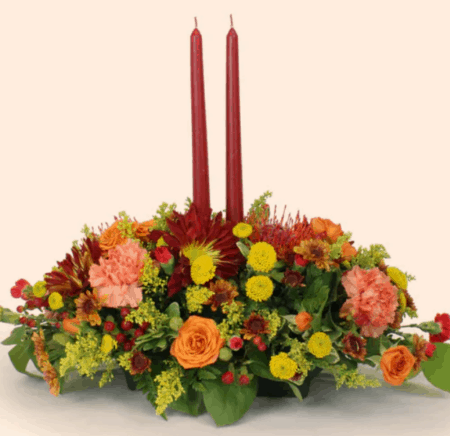 Two taper candles are the center of attention. Perfect for casting that warm holiday glow. The flowers are created in a long compact design so that plenty of conversation can take place over them. Fall flowers such as miniature spray roses and protea are sprinkled among traditional fall chrysanthemums