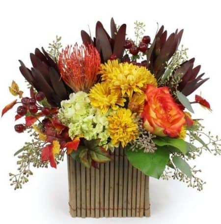 Our Shades of Autumn Bouquet is a wonderful way to celebrate the season. The unique bamboo square container is filled with fall color. We have two varieties of Protea. One in a fiery orange and the other in a bold burgundy. Two tones roses add a splash of elegance. Classic golden Cushion Chrysanthemums add a richness to the design. Miniature green Hydrangeas add a touch of southern charm to the Fall bouquet. Seeded Eucalyptus, Hypericum berries and faux fall leaves add crispness. A perfect way to bring the Shades of Autumn to their home or business.