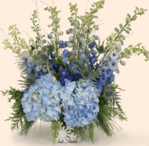 Beautiful blue Hydrangeas are nestled in fragrant evergreens. Blue Delphinium extends upward from the base and is accompanied by silvery blue Thistle. The arrangement is centered in a clear cube embossed with a beautiful snowflake pattern in white.