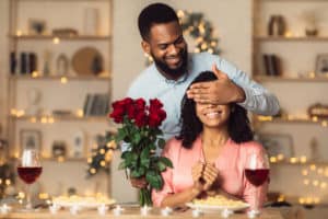 Greeting With Holidays. Smiling black man covering his woman eyes and giving her bunch of red roses, making surprise to beautiful lady. African american couple celebrating together at home or cafe